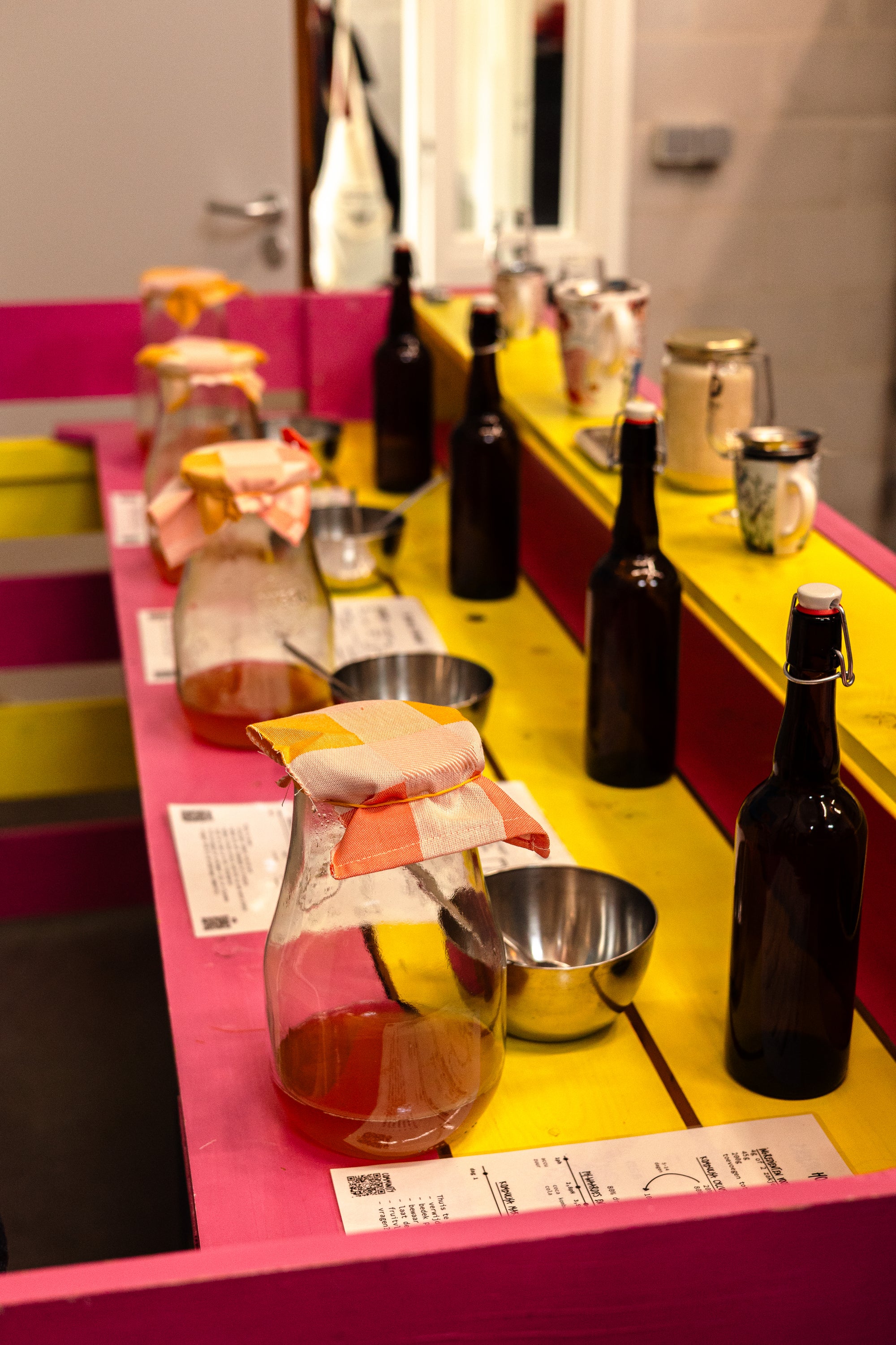 Een tafel met daarop glazen potten met SCOBY, een hand-out met kennis en recepten, en een glazen fles met beugel om kombucha in te doen