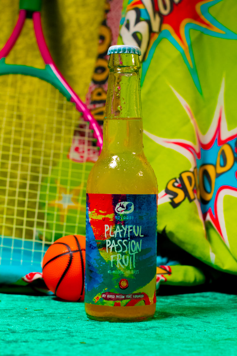 Een flesje Playful Passion Fruit van Untamed Kombucha met een tennisracket op de achtergrond