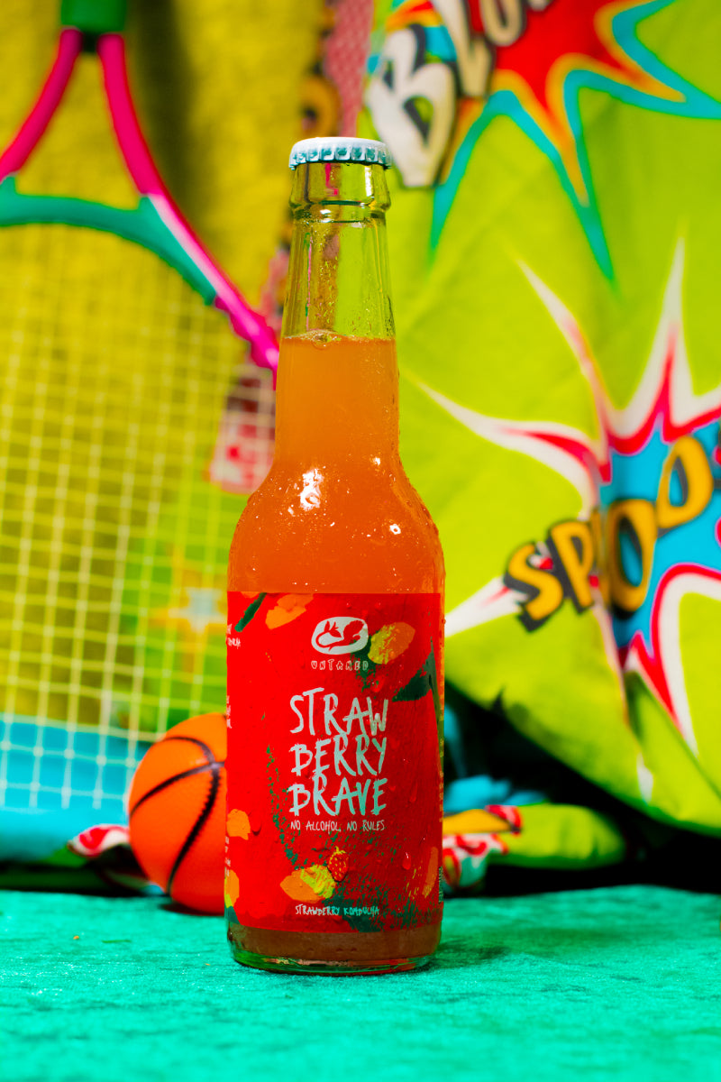 Een flesje Strawberry Brave Untamed Kombucha met een sportieve achtergrond met onder andere een tennisracket
